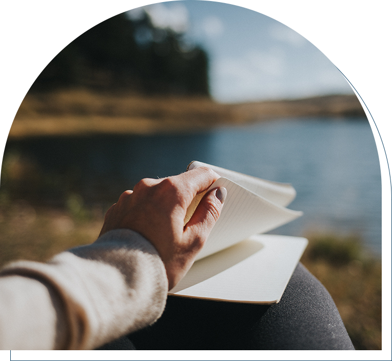 A person writing in a notebook near water.